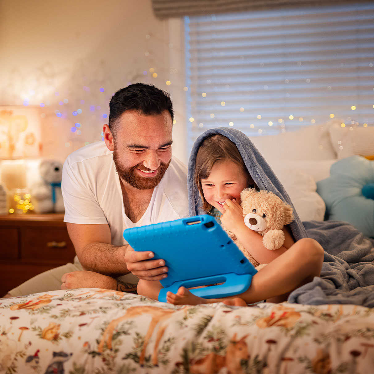 A man and a child sit together on a bed, sharing a tablet under a cozy blanket.